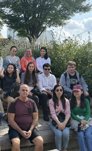 A group of students outdoors sitting in three rows with bushes, a tree, and a fence behind them.