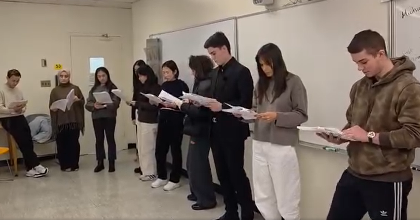 10 diverse students standing in front of a whiteboard looking down at the scripts in their hands.