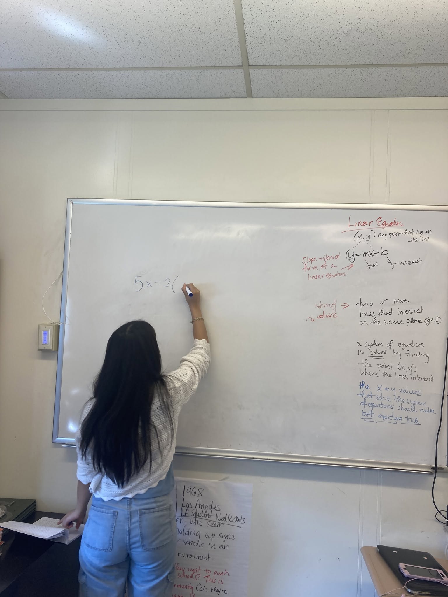 Student in white top and blue jeans with long black hair writing on a whiteboard with a marker