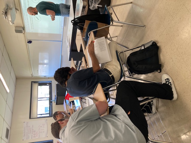 Students in a classroom facing the instructor with class materials projeced onto the whiteboard.