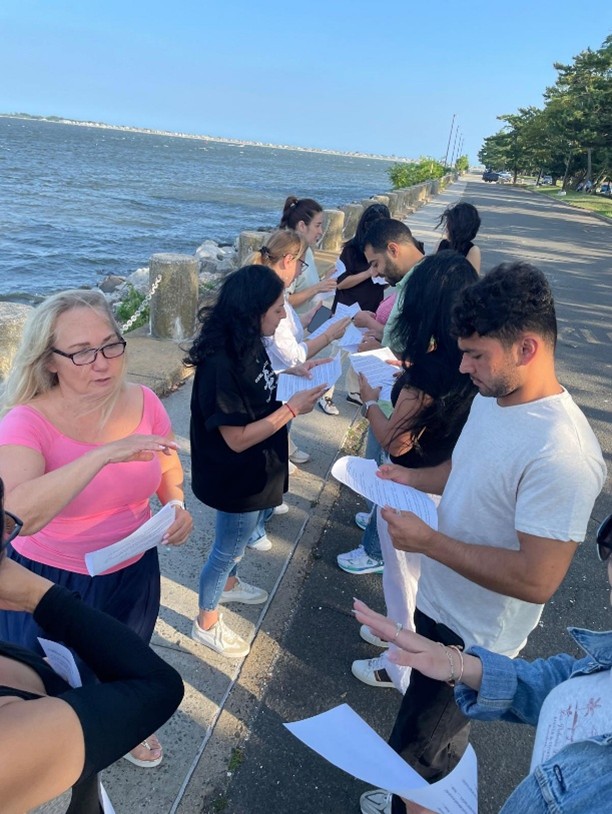 Students on KCC campus outside by the seawall, looking at handouts in their hands and listening to the instructor wearing glasses and a pink top. 